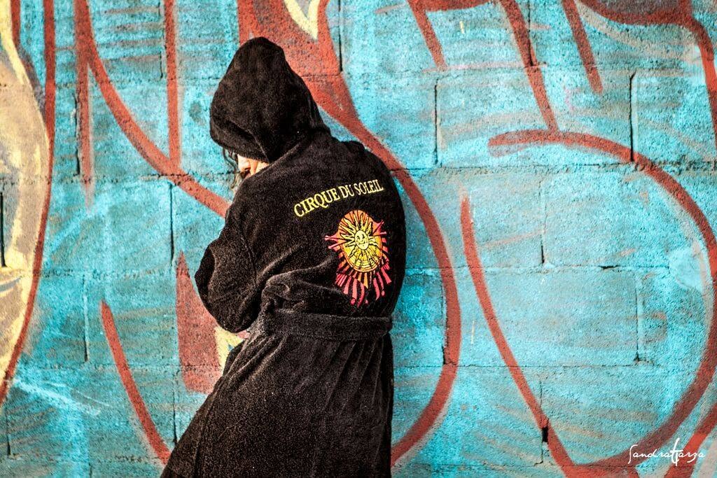 Person in black hoodie with a colorful sun design on back, standing against a graffiti-covered wall.