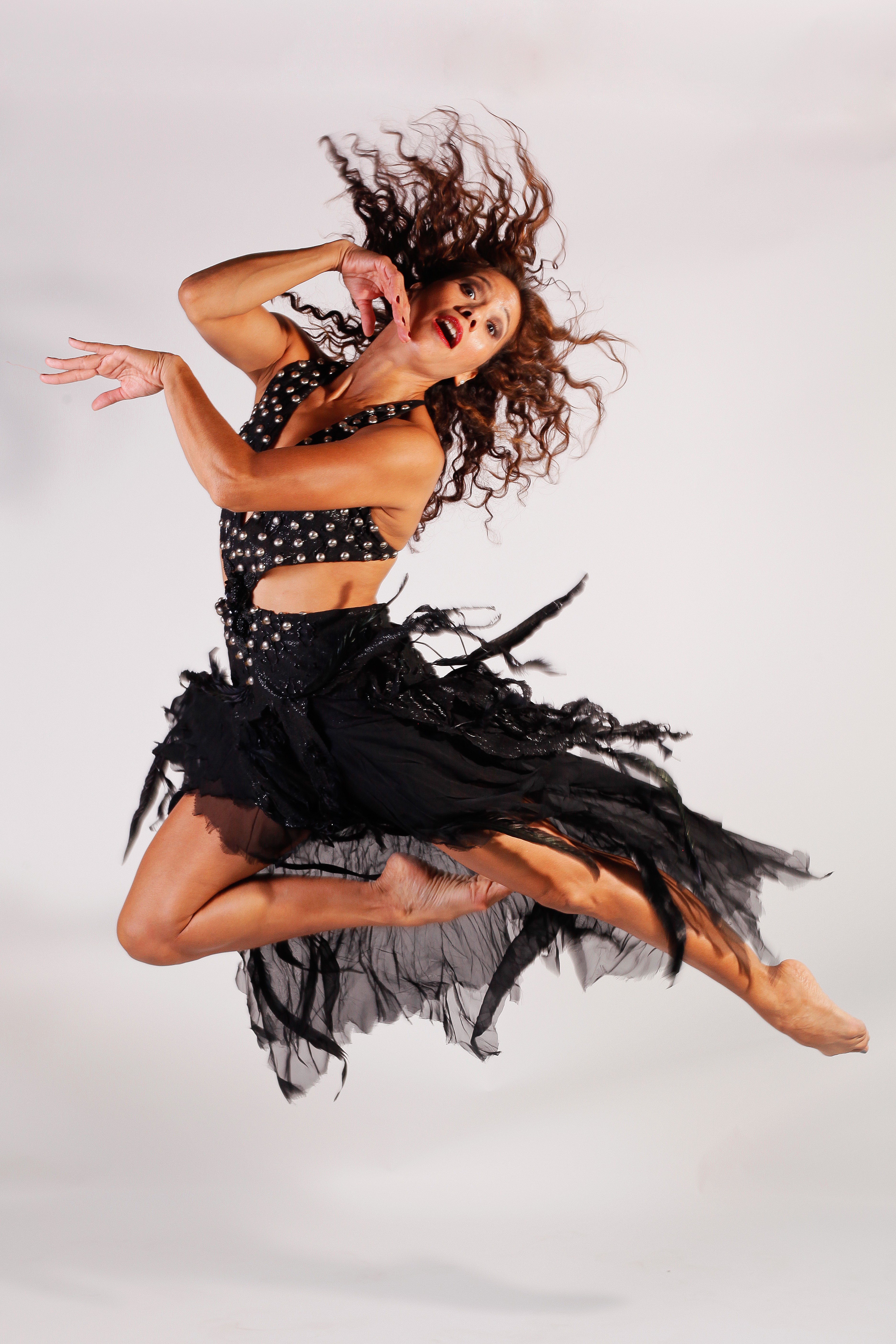 A woman with curly hair jumps mid-air wearing a black polka dot top and a black skirt, against a plain background.