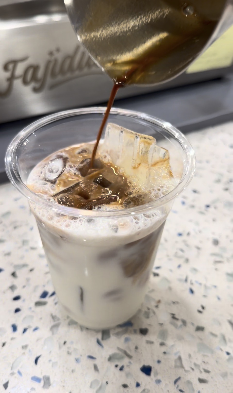 Coffee is poured into a glass with ice, creating a layered drink on a speckled surface.