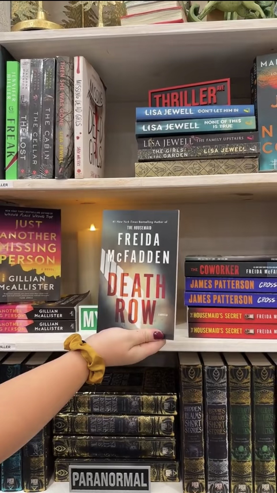 Person's hand holding a book titled 'Death Row' in front of a bookshelf with various books, some with glowing lights.