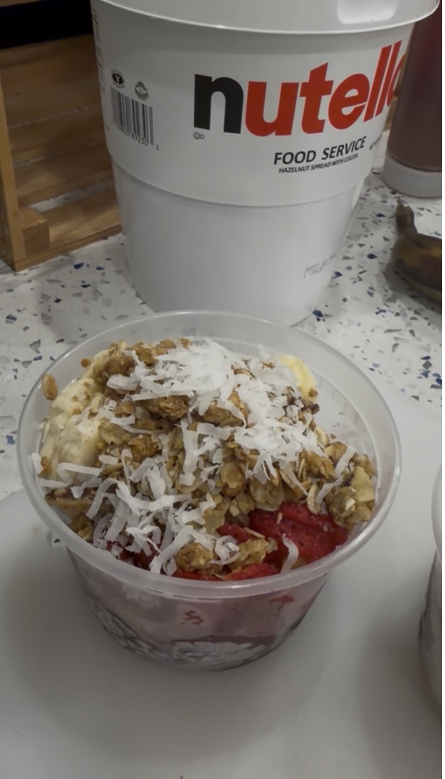 Bowl of layered dessert with crushed cookies, coconut flakes, and fruit on top, on a speckled countertop.