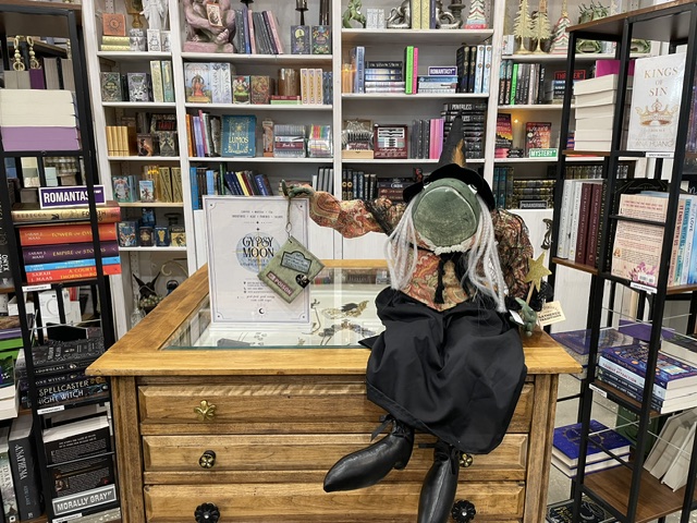 Person with dreadlocks wearing headphones, sitting at a wooden table in a bookstore, facing shelves filled with books.