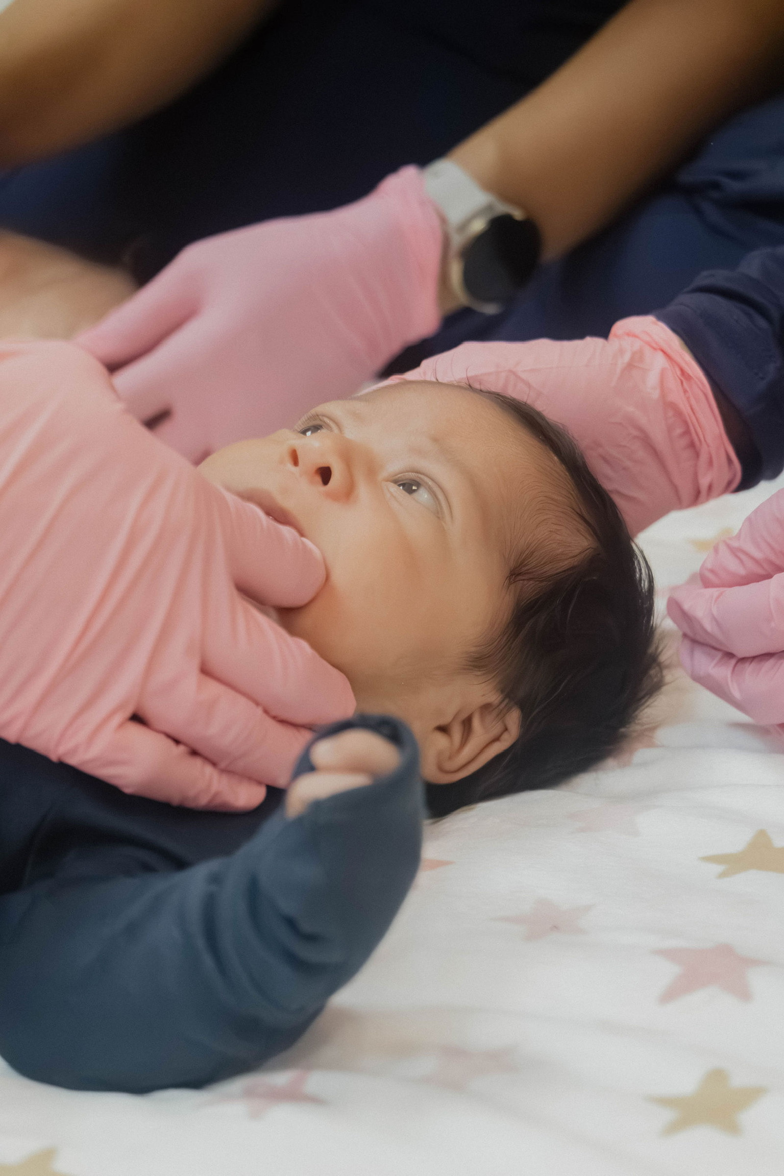 Baby lying on bed with a pink blanket, looking up, person in pink gloves touching face, star-patterned sheet visible.