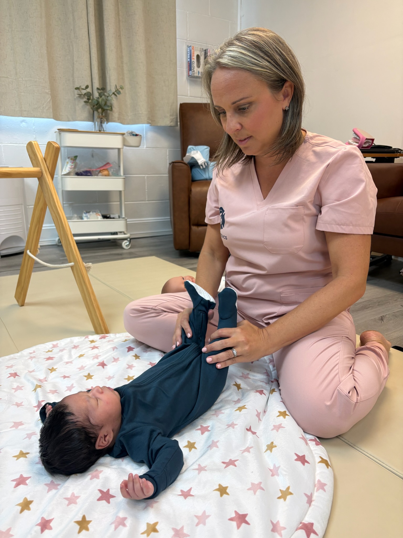 Woman in pink scrubs kneels on floor with a baby lying on a star-patterned blanket, holding the baby's legs.