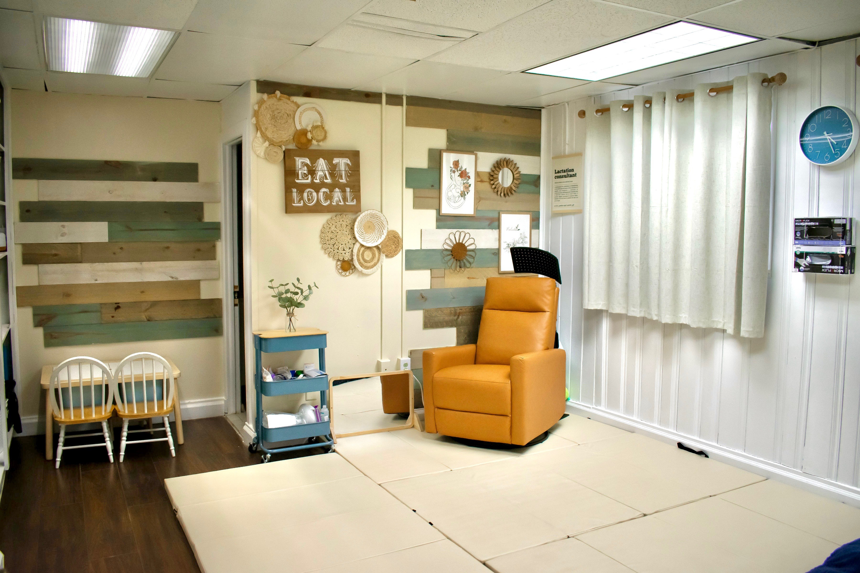 Room with a yellow armchair, wall decorations, small chairs, and a cart, with white curtains and wall paneling.