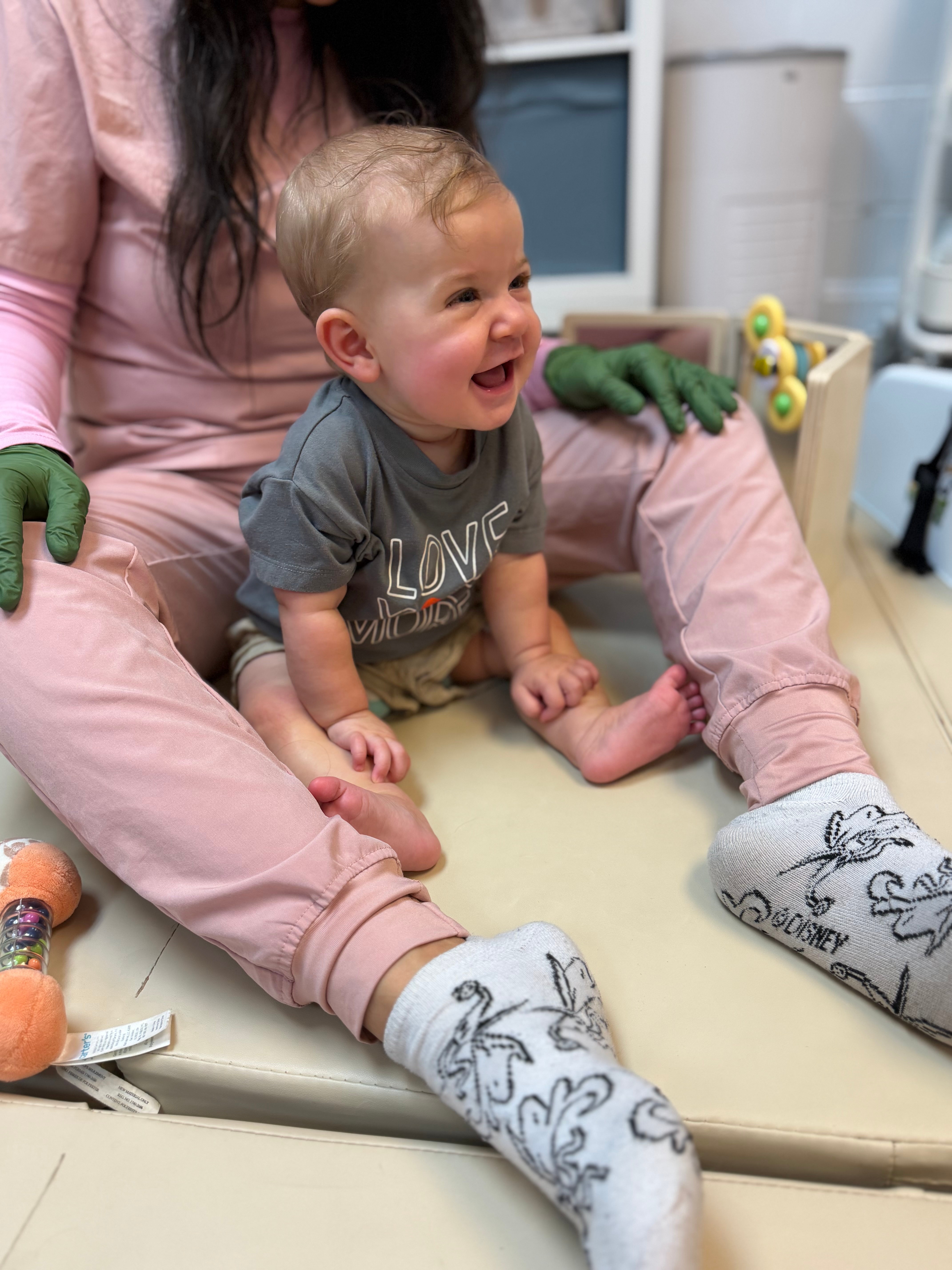 Child sitting on a padded surface, smiling, with a person in pink clothing and green gloves nearby, in a room with toys.