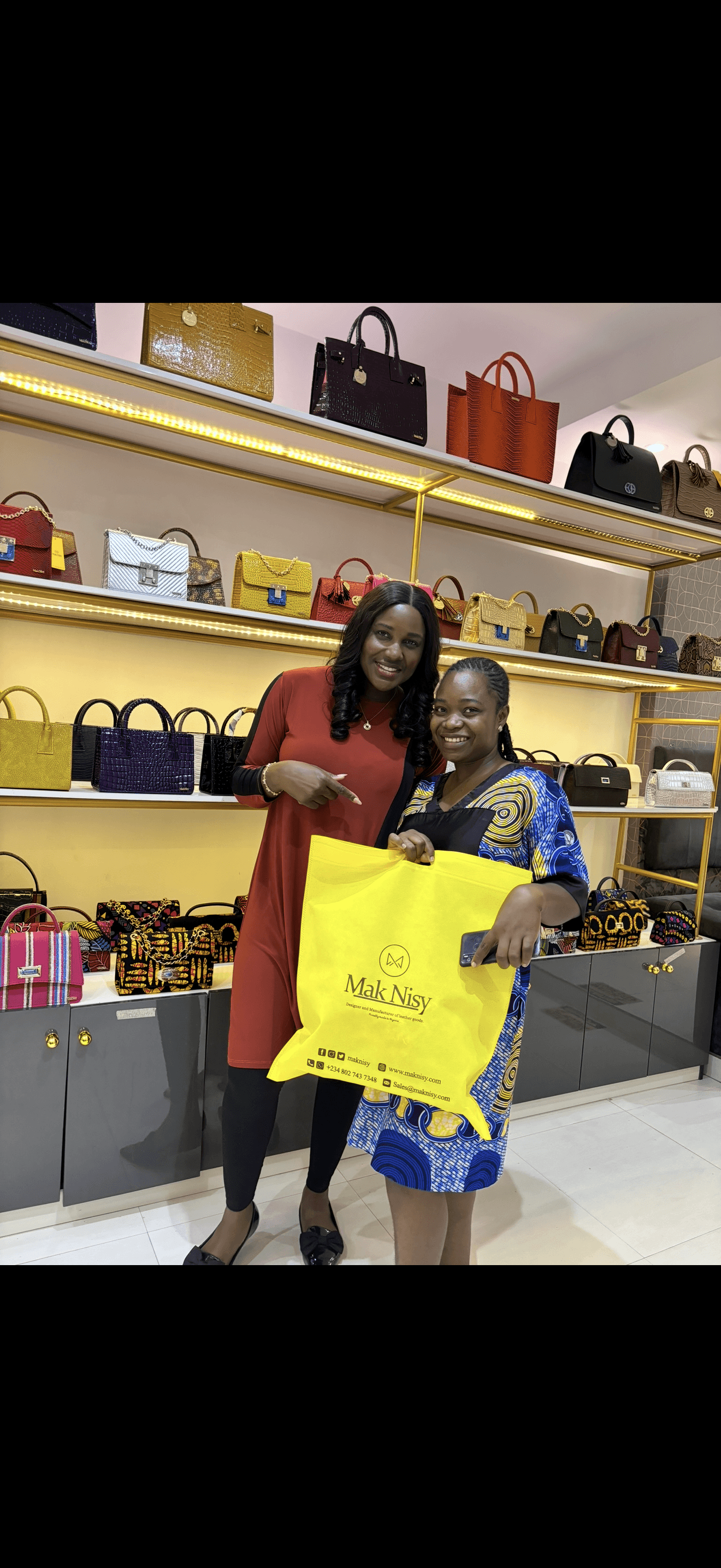Two women shopping for handbags in a store, holding a yellow shopping bag, shelves with handbags behind them.