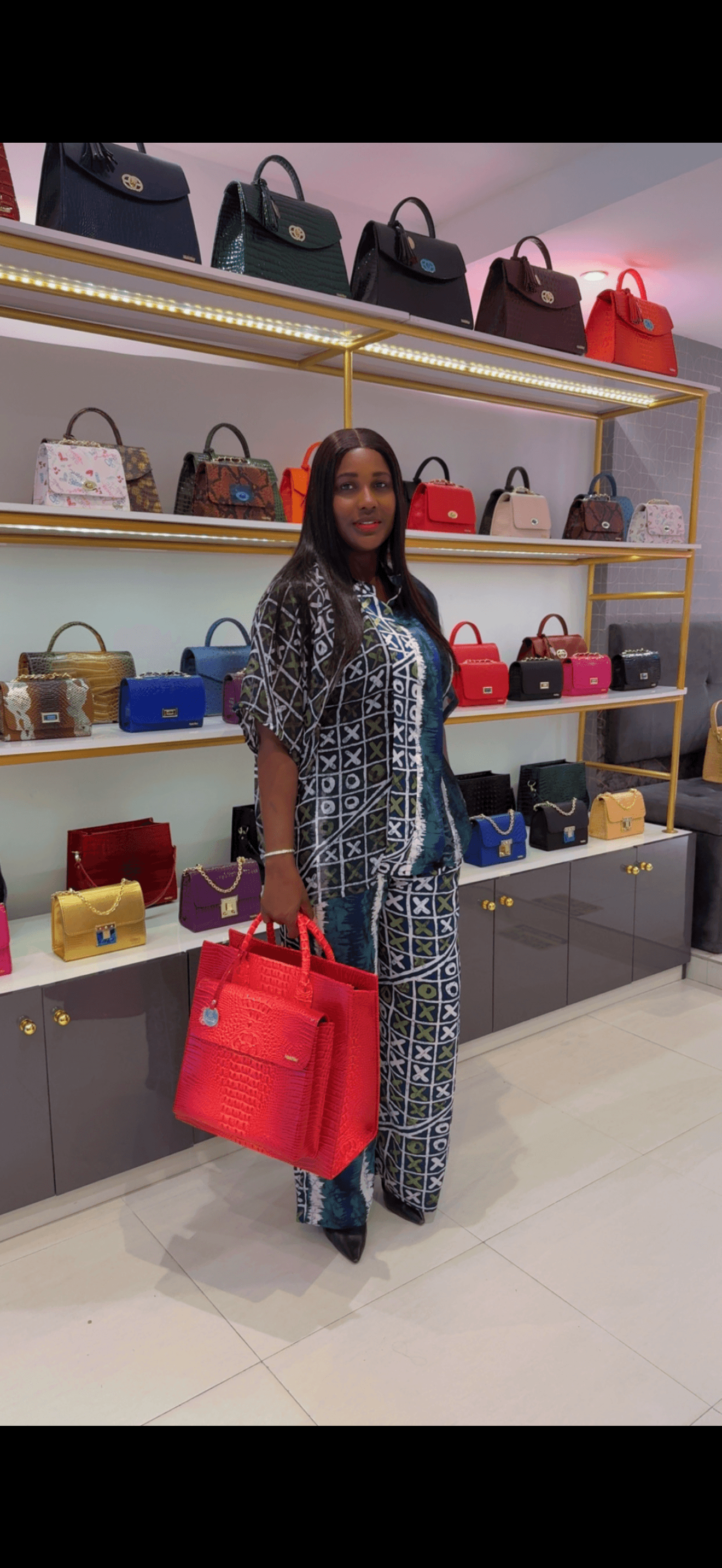 Woman in patterned outfit holding a red handbag in a store with shelves of handbags behind her.