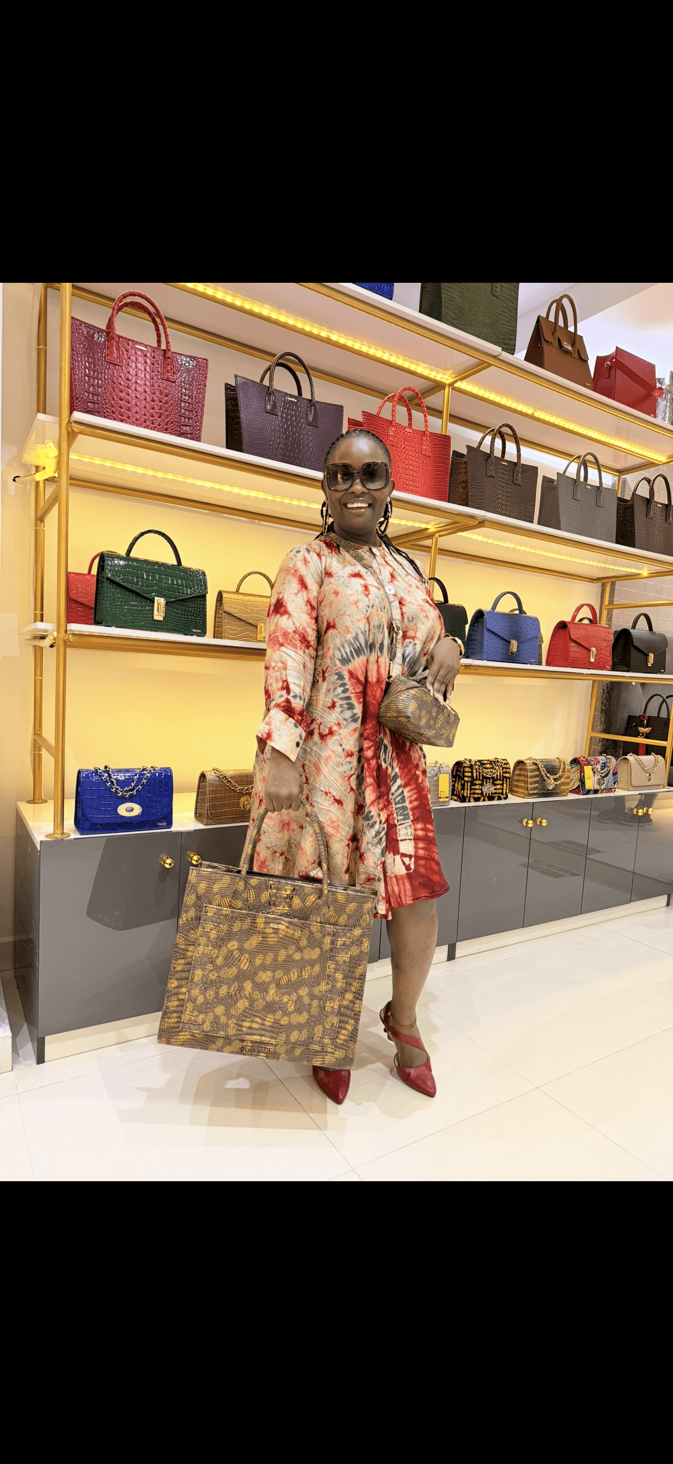 Person standing in front of shelves filled with handbags, holding a large patterned bag, wearing a floral top and red skirt.