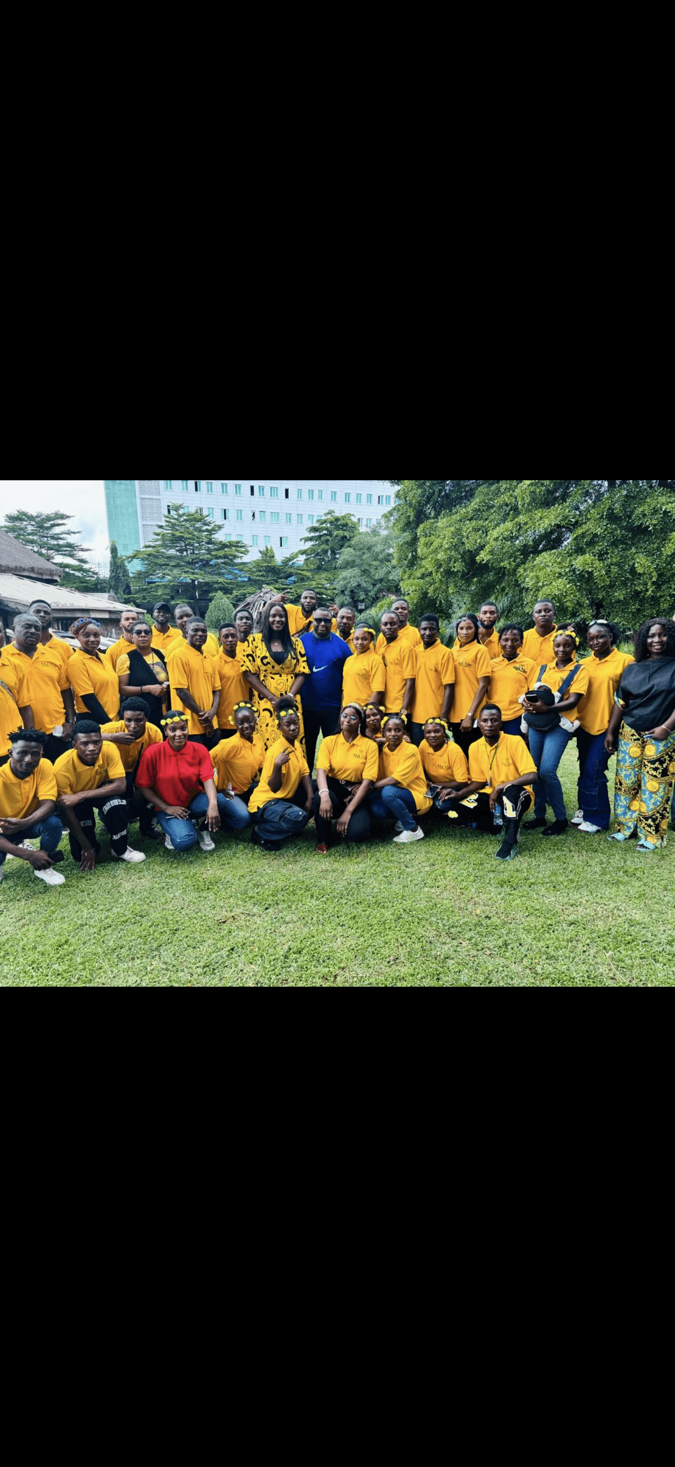 Group of people outdoors, some in yellow uniforms, others in casual clothing, with trees and buildings in background.