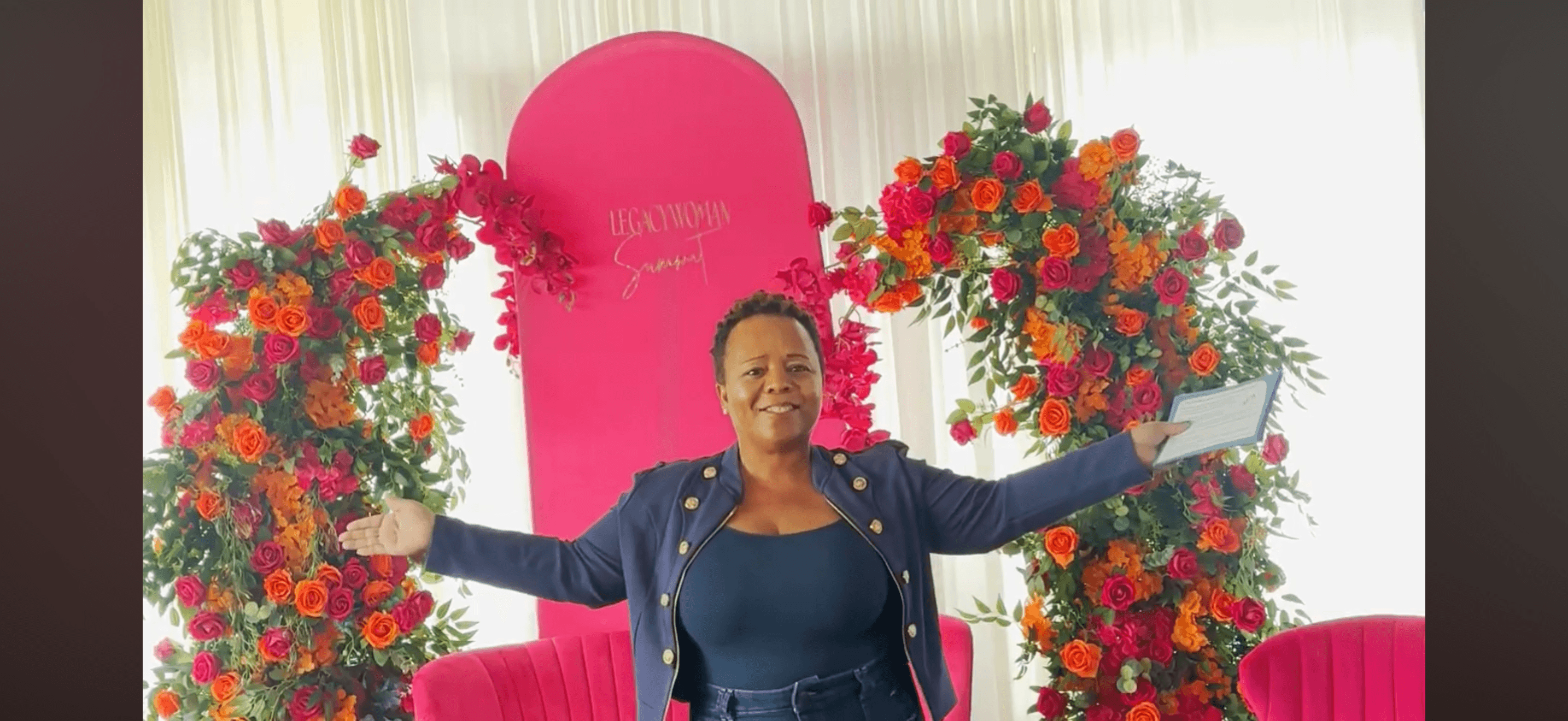 Woman standing with arms outstretched in front of floral arrangements and a pink backdrop, smiling.