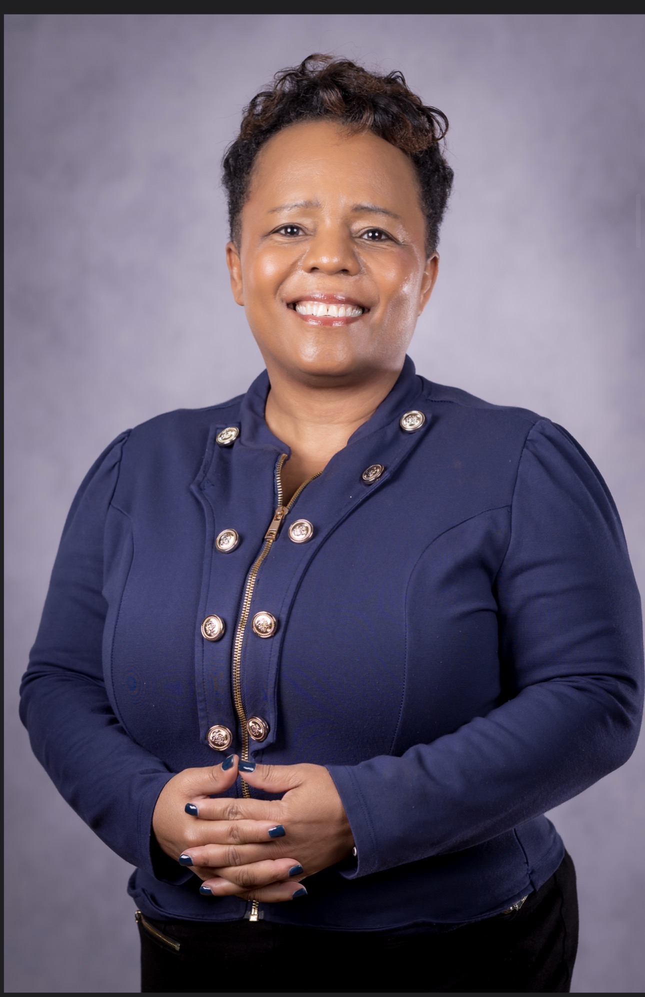 Smiling woman with short curly hair wearing a navy blue jacket with gold buttons, standing against a gray background.
