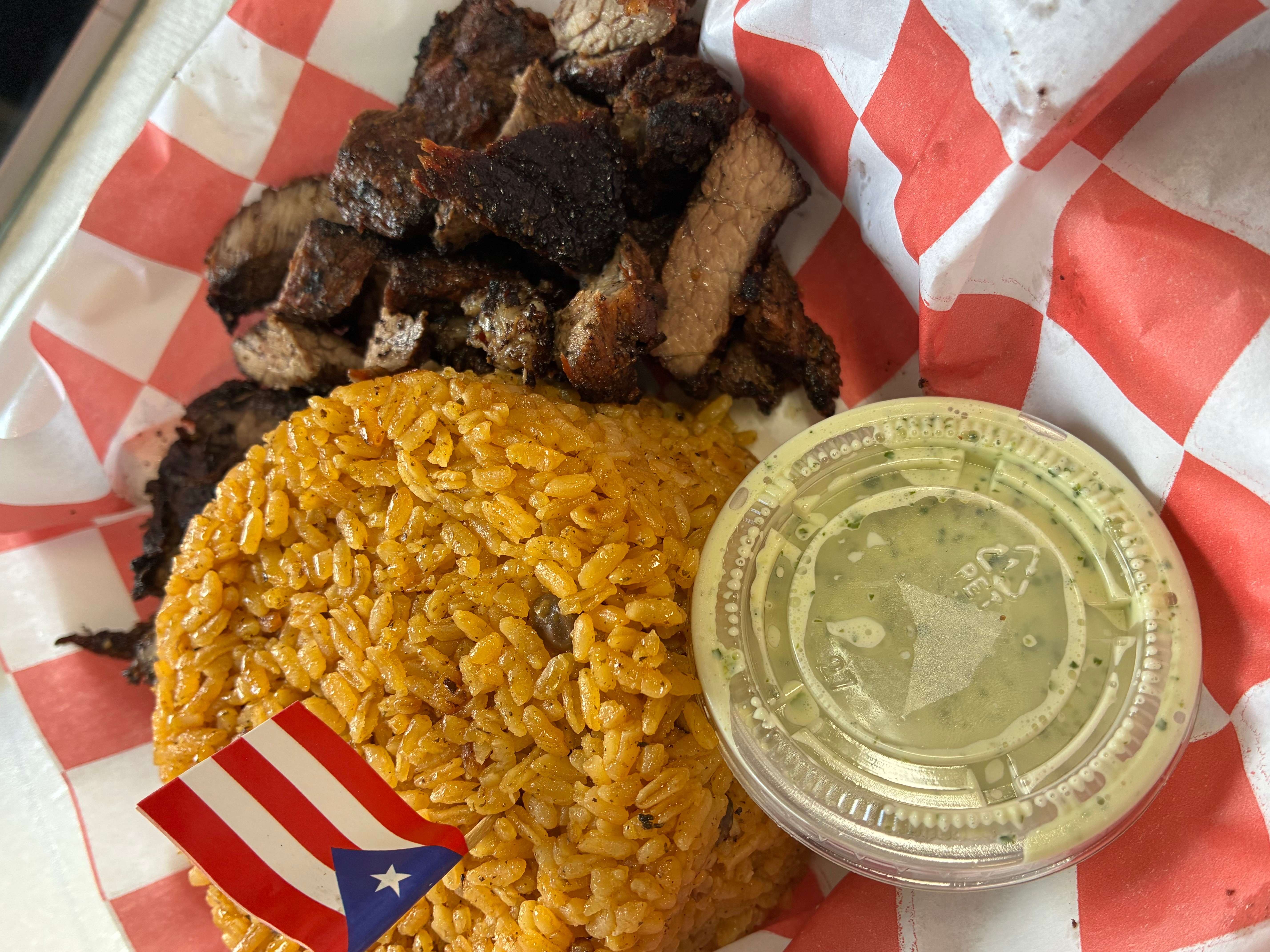 Plate with yellow rice, fried meat pieces, and a small container of sauce on red and white checkered paper.