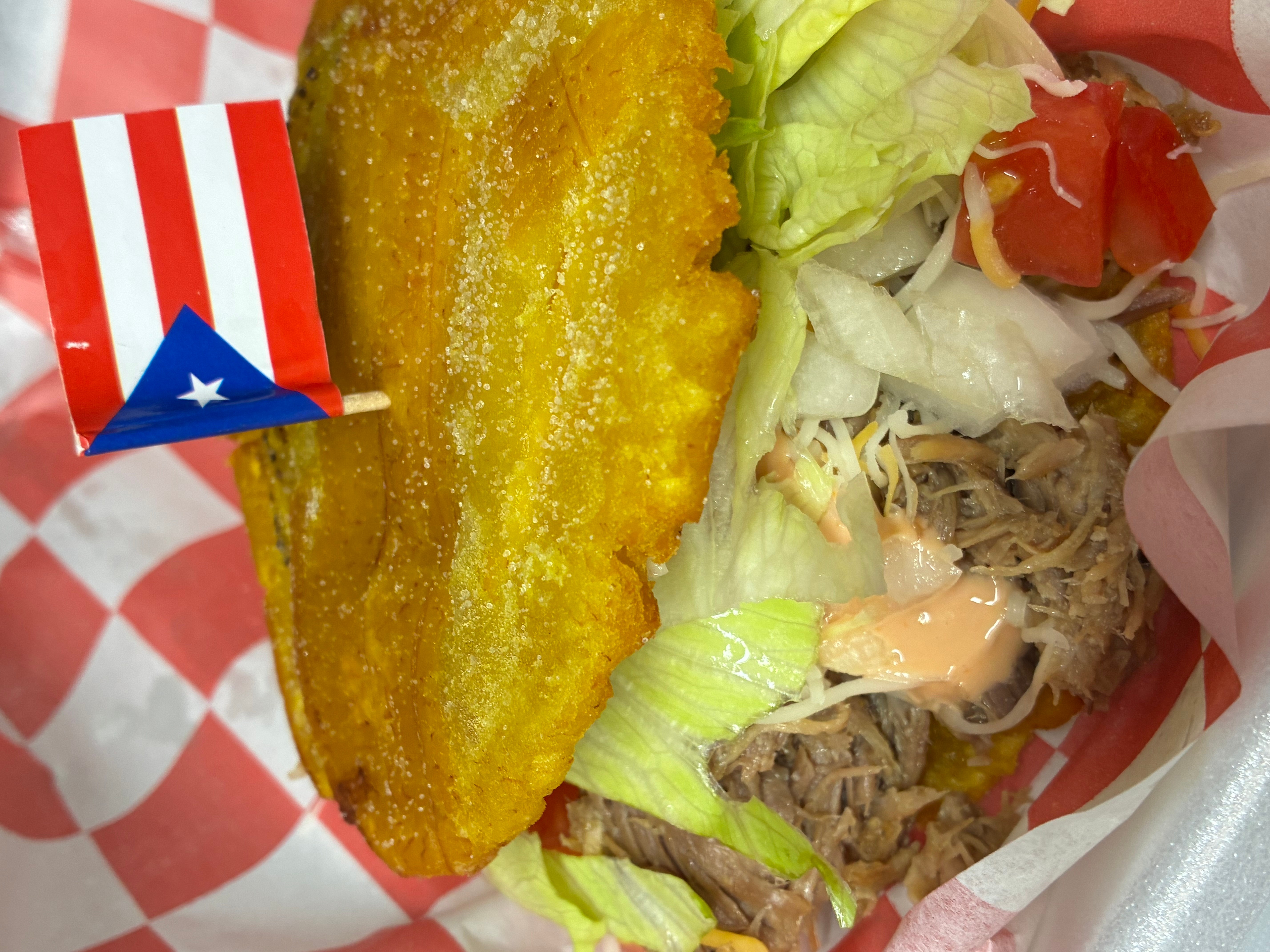 Fried fish fillet with lettuce, shredded meat, and tomato in a basket with checkered paper, and a small Puerto Rican flag decoration.