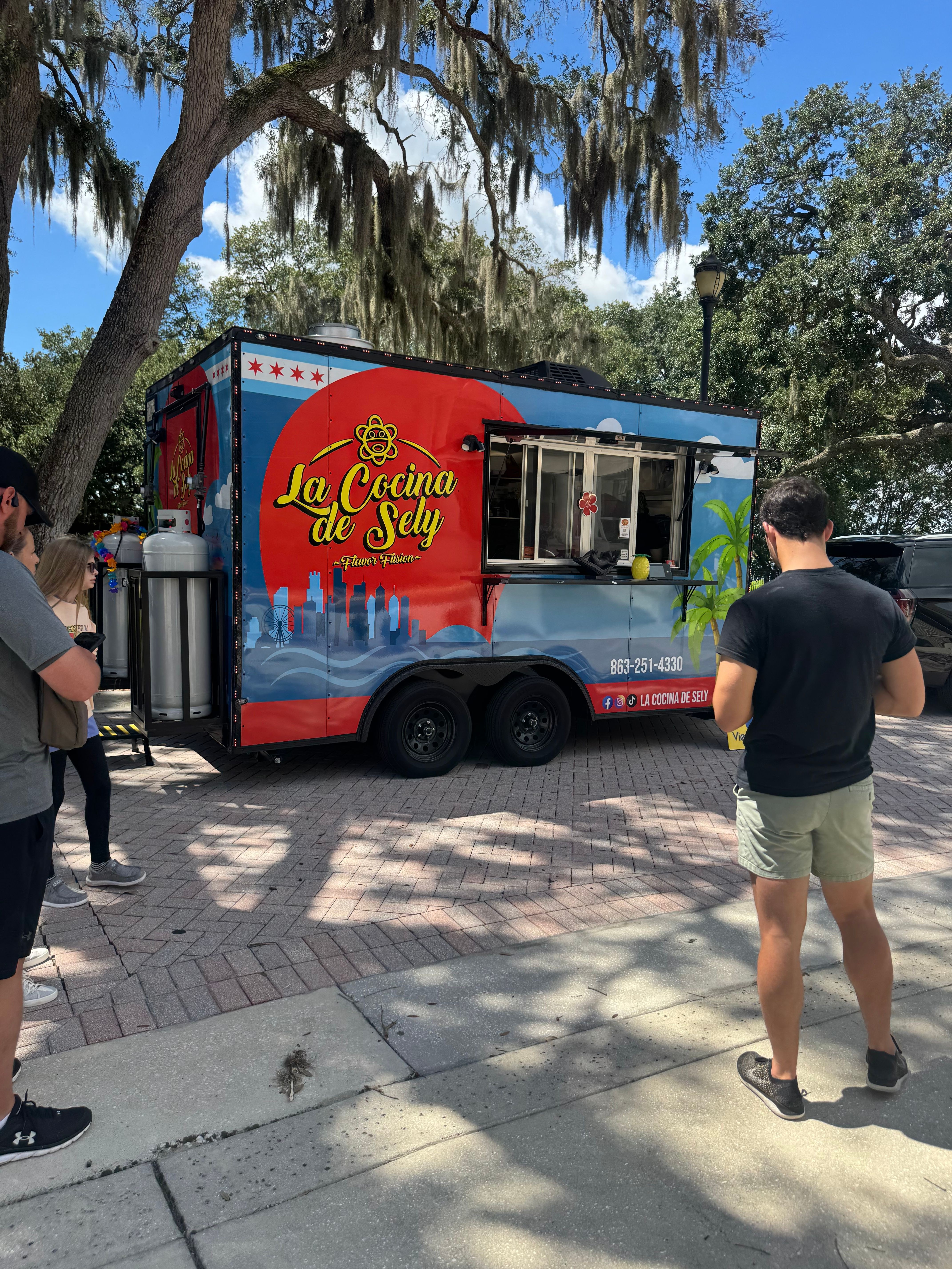 Ice cream truck with colorful design parked on sidewalk, people walking nearby, trees and blue sky in background.