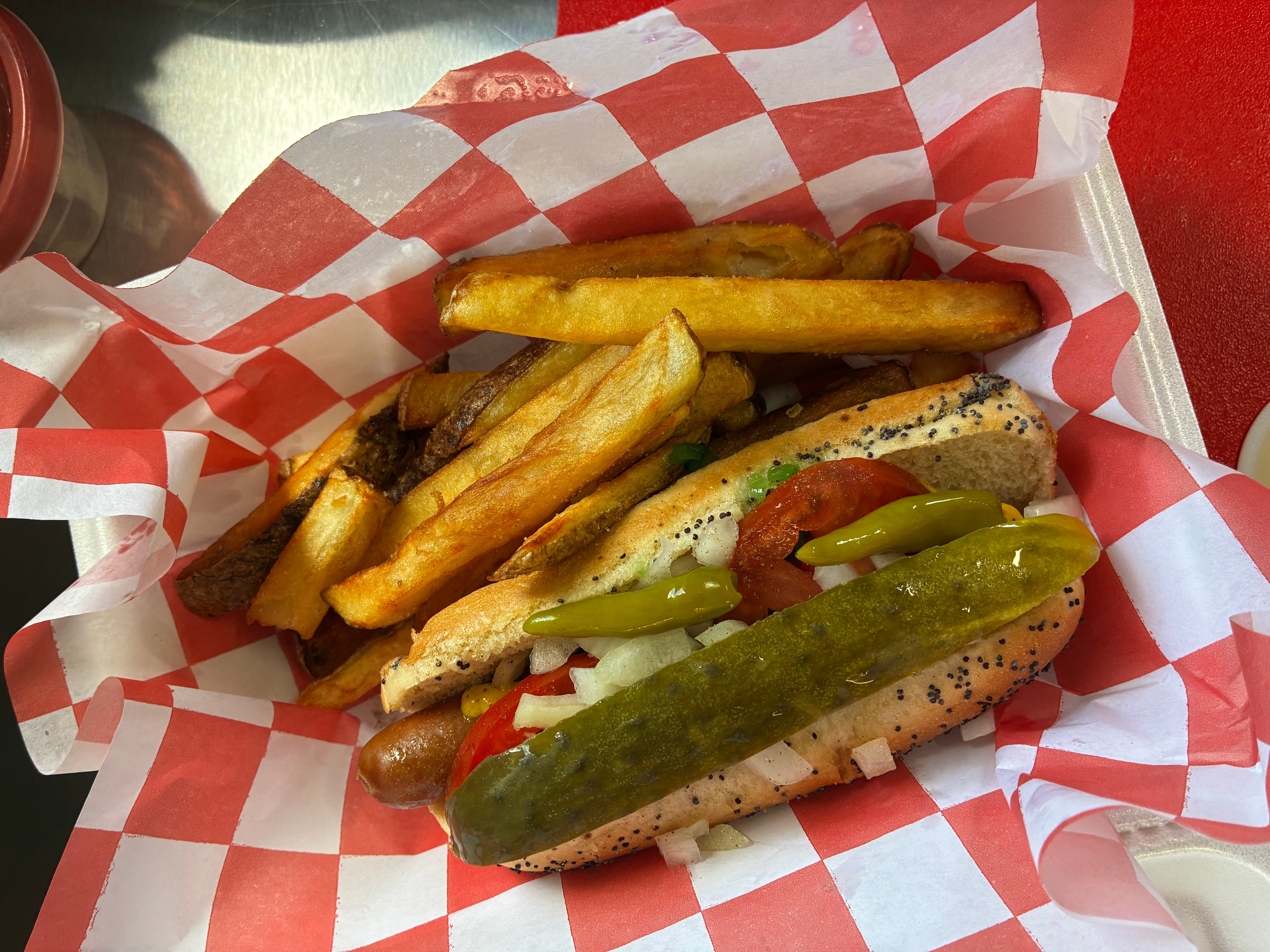 Hot dog with mustard, pickles, and poppy seed bun, served with French fries on red and white checkered paper.