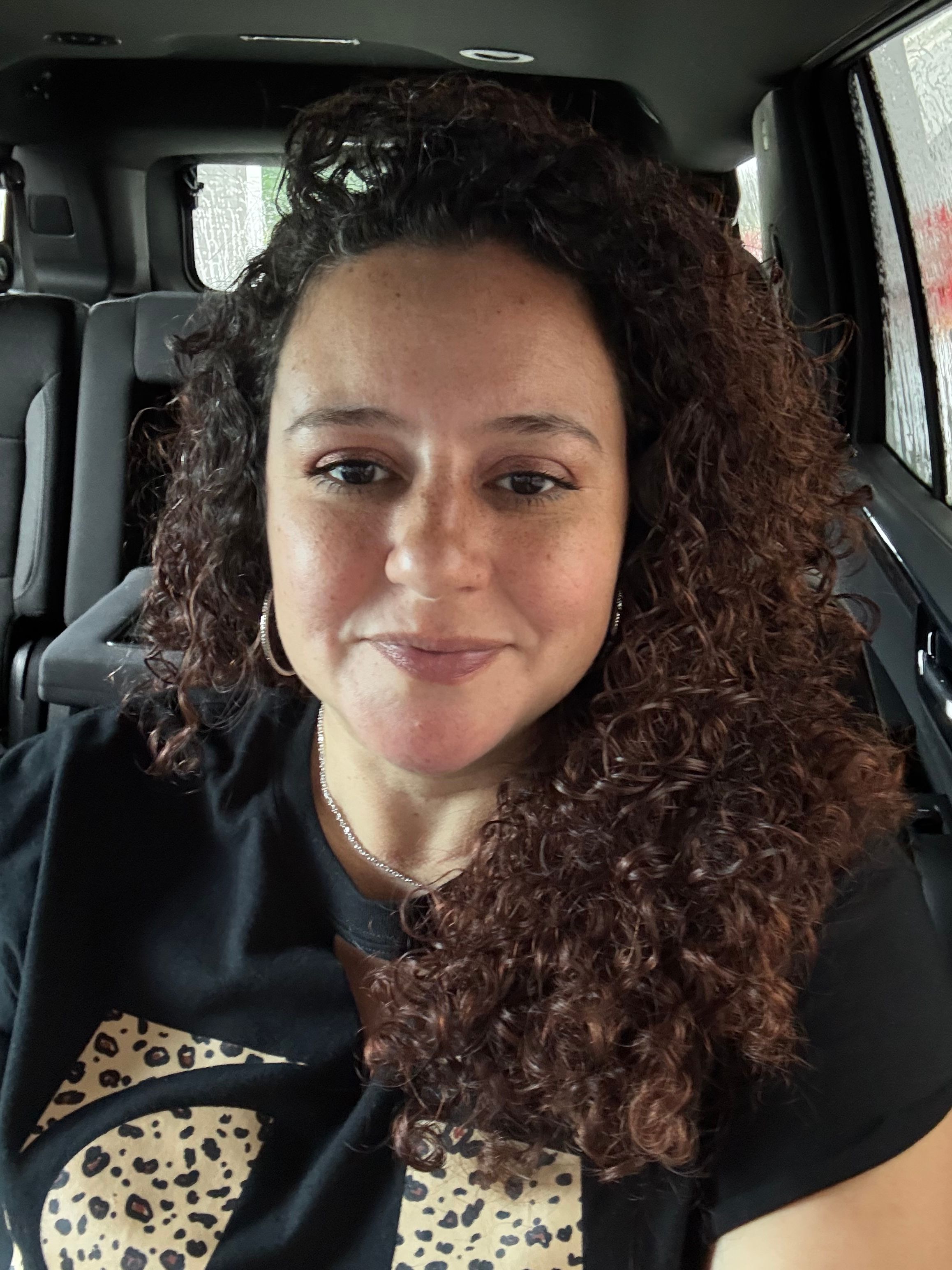 Woman with curly hair sitting in a vehicle, smiling, wearing a dark top with a patterned detail.