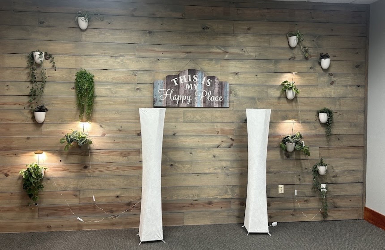 Decorative wooden wall with potted plants and a sign, two tall white planters in front, and small wall lights.