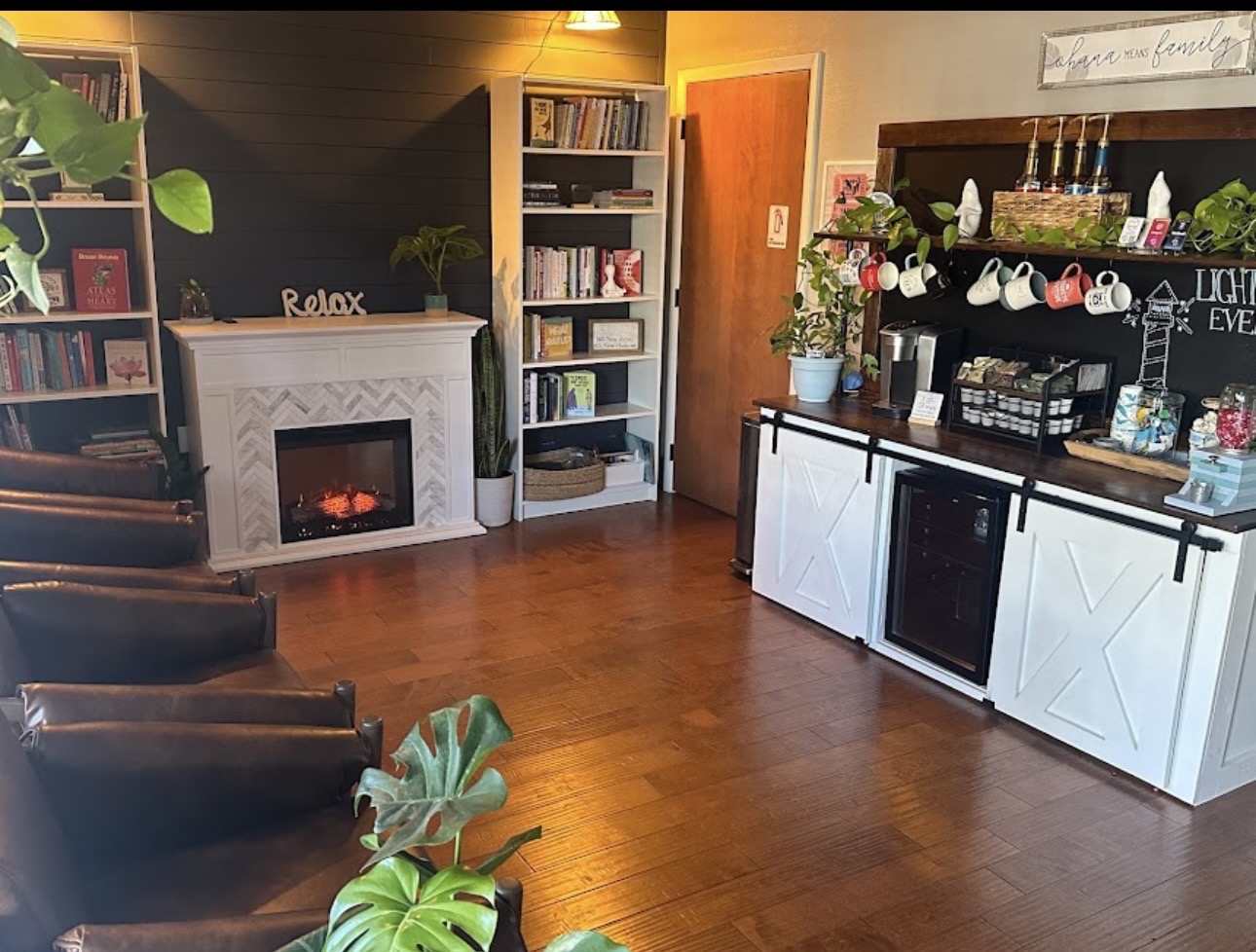 Cozy room with chairs, bookshelf, fireplace, and a bar area with decorations and plants, warm lighting, and wooden flooring.