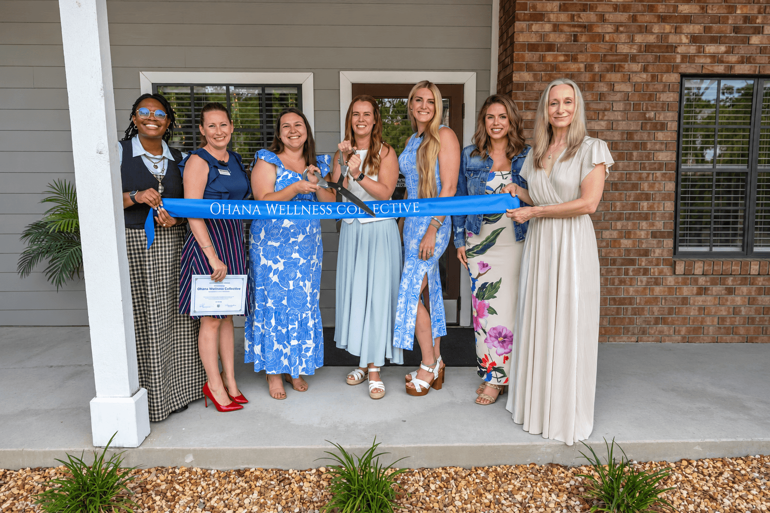 Group of seven women standing outside, holding a blue ribbon with text, smiling, in front of a building with brick and siding.