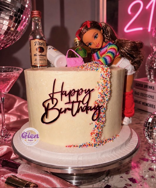 A birthday cake with a doll on top, colorful sprinkles, and a bottle of liquor, surrounded by party decorations.