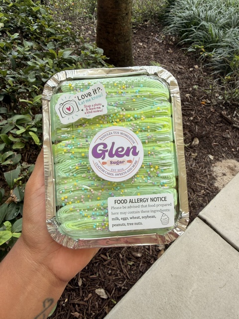 Hand holding a container of green and yellow layered food outdoors, with plants and concrete nearby.