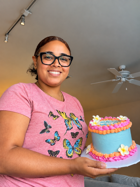 Smiling woman with glasses holding a decorated cake, wearing a pink butterfly-printed shirt, ceiling fan above.