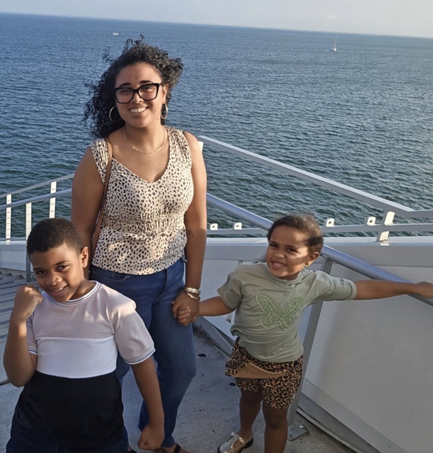 Woman with curly hair and glasses holding hands with two children on a boat, ocean in background.