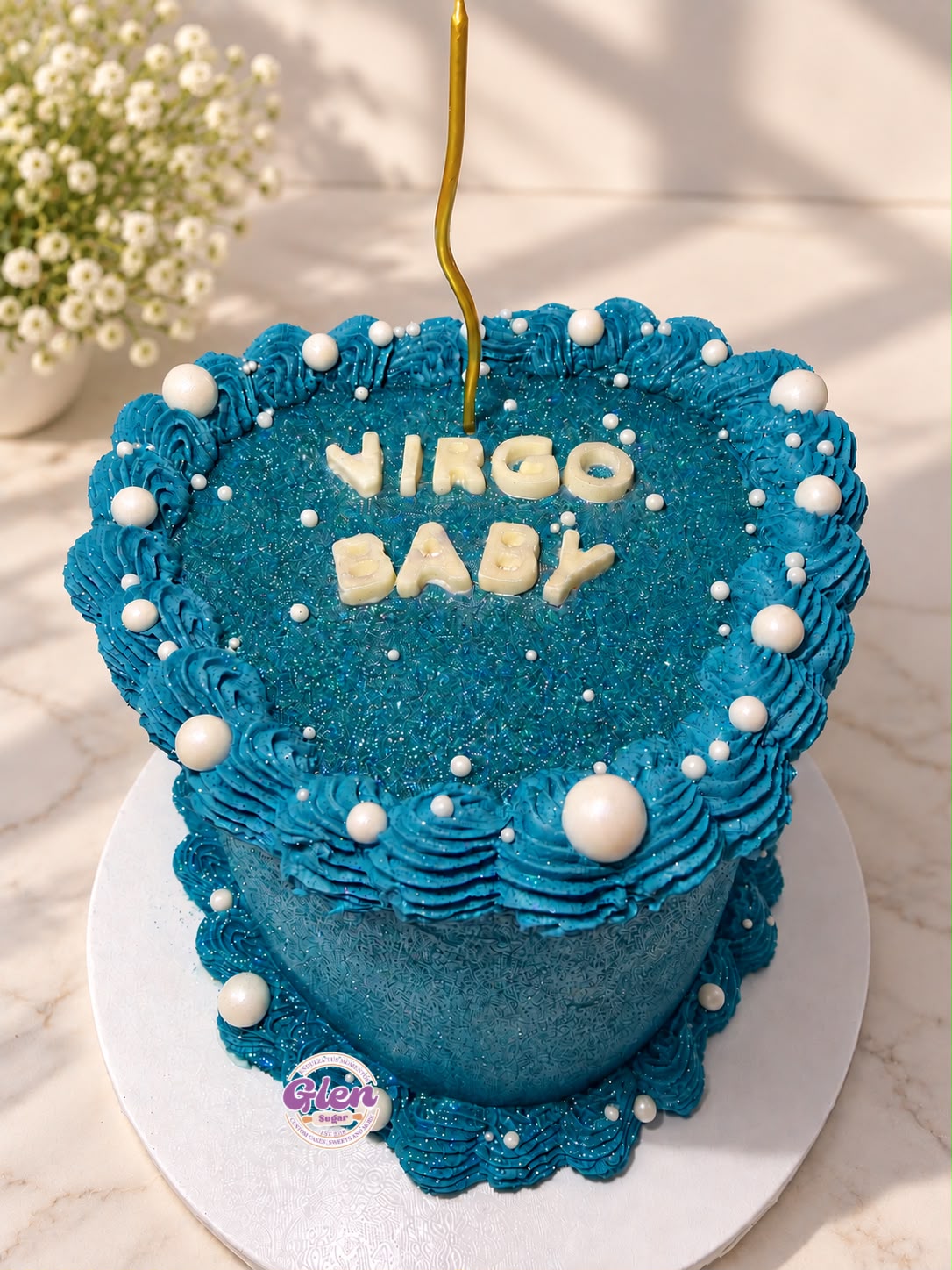 Heart-shaped birthday cake with blue frosting, white decorative balls, and the words 'Virgo Baby' on top, with a candle.