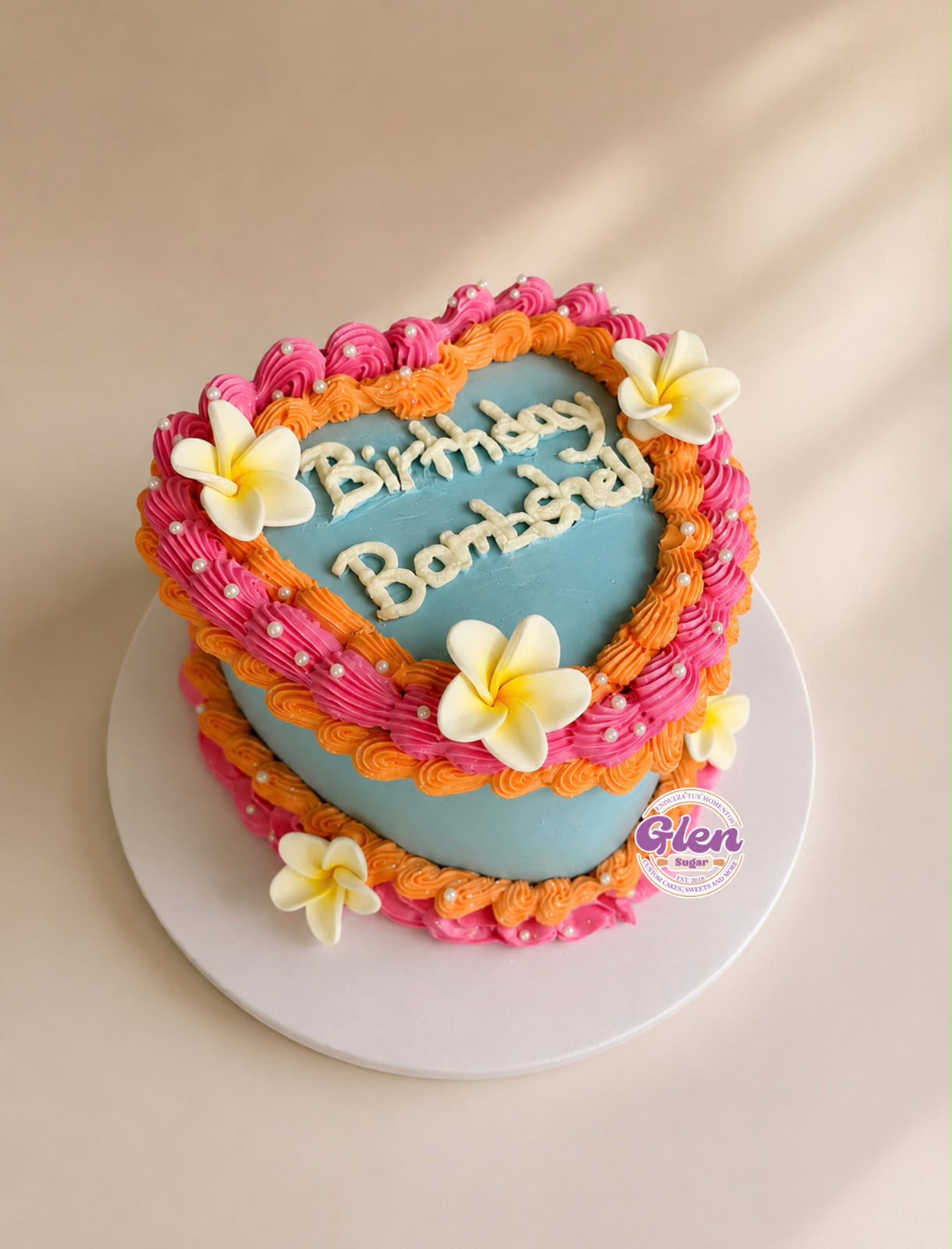 Heart-shaped birthday cake decorated with pink, orange, and white flowers, with blue icing and birthday message, on a white surface.