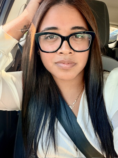 Young woman with glasses and long hair, sitting in a car, looking at the camera.