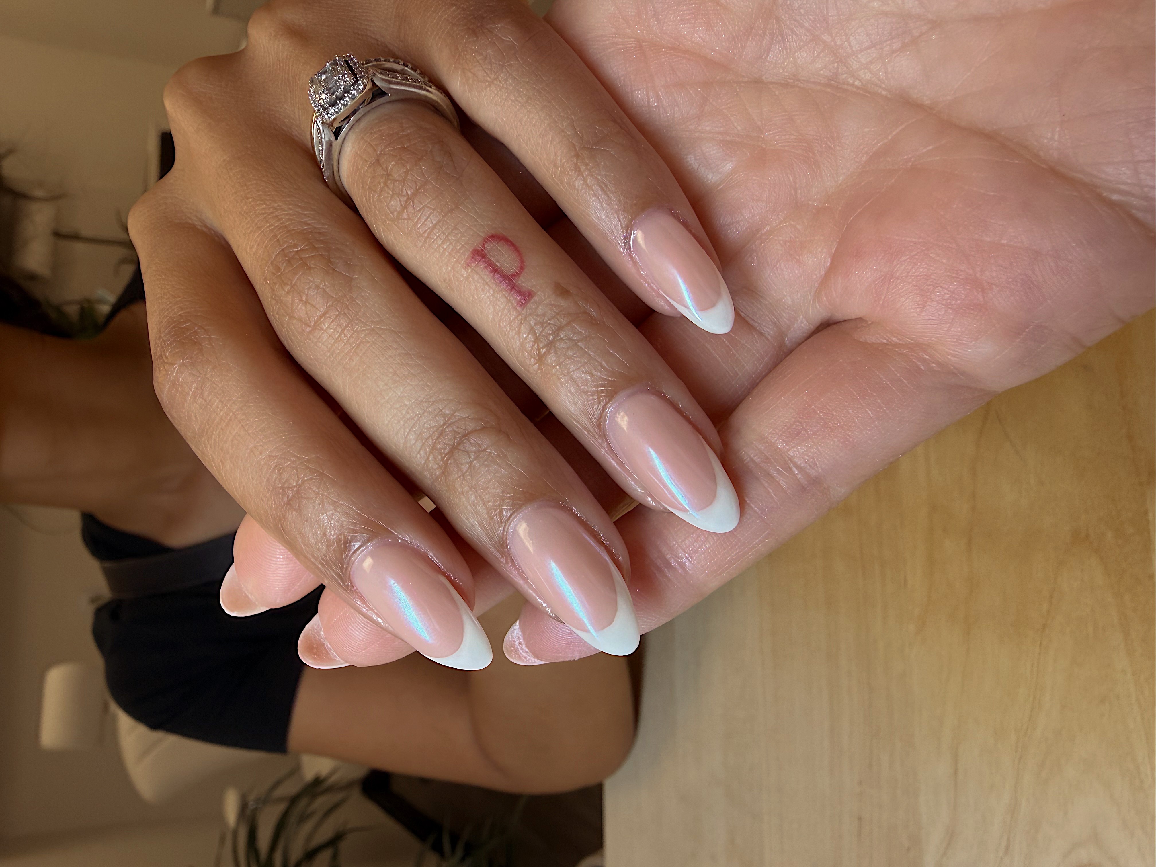 Close-up of a hand with manicured nails resting on a wooden surface, showing rings and a tattoo on the finger.