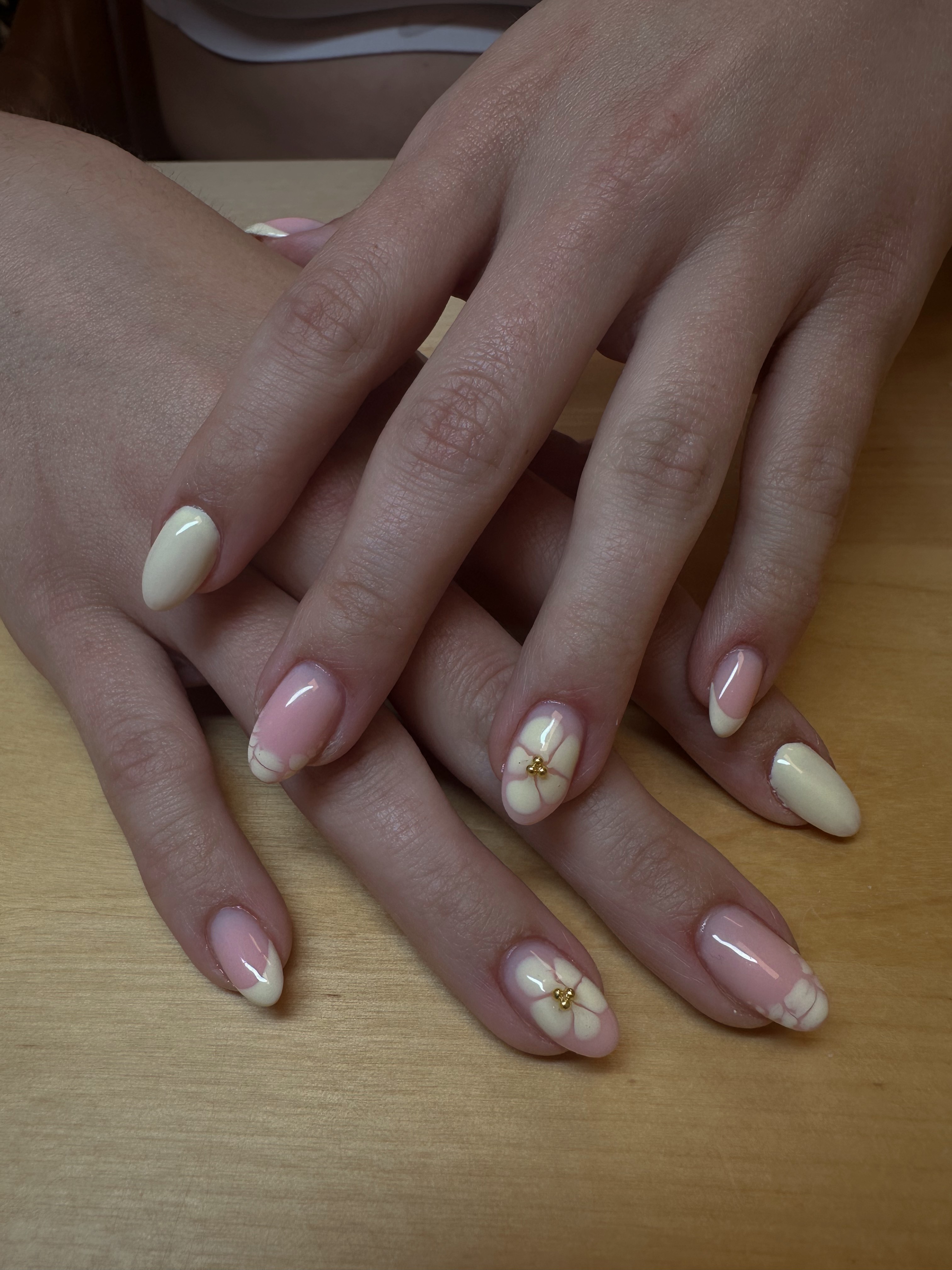Two hands with manicured nails resting on a wooden surface, one hand with white and gold floral nail art, the other with plain nails.